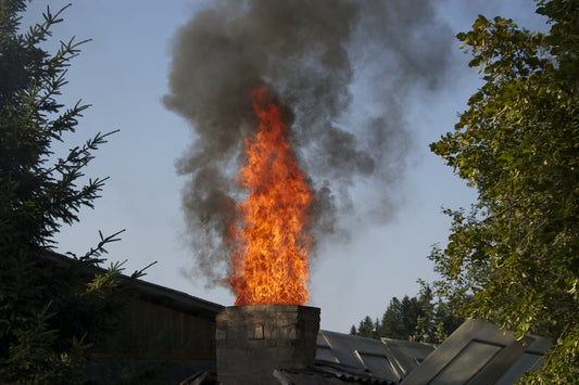 Chimney Fire. A large, orange flame erupts from a chimney, with thick, dark smoke rising above it. The fire is set against a blue sky and green trees.