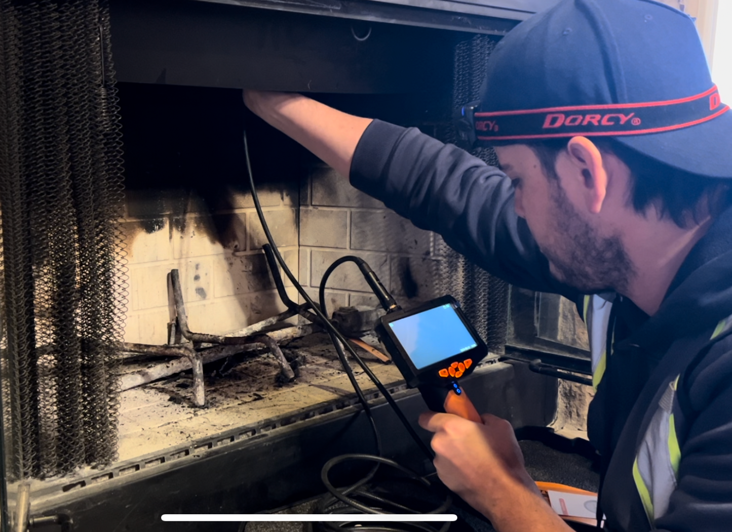 A person wearing a blue baseball cap with a red headlamp and a black face mask is inspecting the inside of a fireplace using a small camera with a screen. The fireplace interior is dark and shows signs of soot and ash buildup on the brick and grate. The person is holding the camera in their right hand and appears to be carefully examining the firebox.