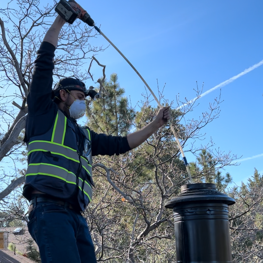 A person wearing safety gear, including a reflective vest and face mask, stands on a roof and uses a power drill to operate a chimney cleaning brush inserted into a black chimney flue against a clear blue sky.